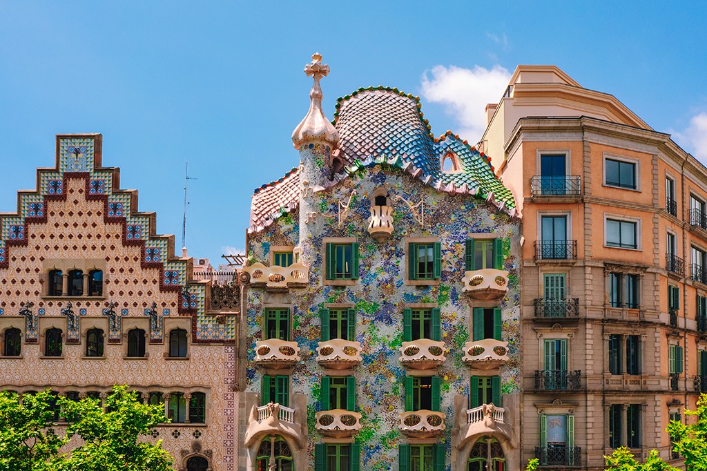 Tr&ecirc;s edif&iacute;cios de arquitetura distinta sob c&eacute;u azul. &Agrave; esquerda, um pr&eacute;dio com fachada em tons de bege e marrom, com telhado em degraus e detalhes em mosaico azul. No centro, a Casa Batll&oacute; de Gaud&iacute;, com fachada colorida em mosaicos, telhado ondulado e chamin&eacute;s decoradas. &Agrave; direita, um edif&iacute;cio cl&aacute;ssico de cor p&ecirc;ssego com sacadas de ferro forjado