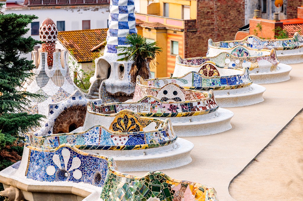 Bancos ondulados de concreto branco adornados com mosaicos coloridos em tons de azul, verde, amarelo e marrom, em um parque com arquitetura modernista ao fundo