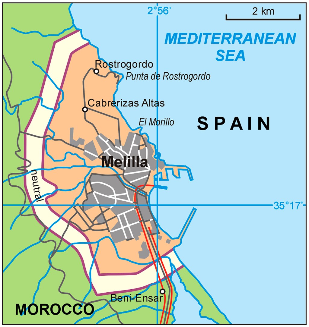Mapa de Melilla, cidade aut&ocirc;noma espanhola na costa do Mar Mediterr&acirc;neo, cercada por Marrocos. A &aacute;rea urbana cinza destaca-se na pen&iacute;nsula laranja, com estradas e o porto. A fronteira com Marrocos &eacute; marcada por uma linha roxa, e uma &aacute;rea neutra clara separa os pa&iacute;ses. Coordenadas geogr&aacute;ficas e escala de 2 km est&atilde;o vis&iacute;veis
