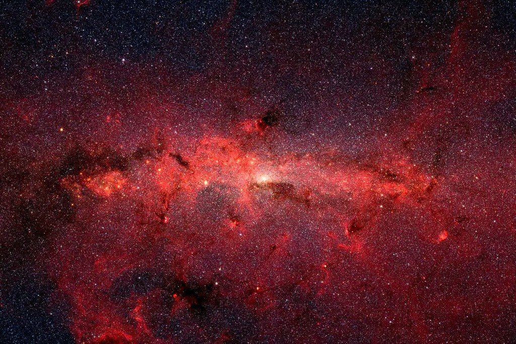 Vasta imagem do centro da Via Láctea, com nuvens de gás e poeira em tons de vermelho intenso e laranja, pontilhadas por milhões de estrelas brancas e amarelas, algumas mais brilhantes, contra um fundo escuro do espaço