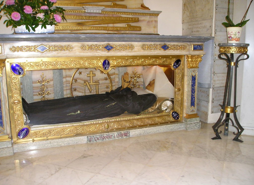 Corpo incorrupto de Santa Catarina Labouré, vestida com hábito preto e touca branca, em um relicário dourado e de vidro, com detalhes em azul e mármore. Ao fundo, uma parede com placas de mármore e, à direita, um pedestal de ferro forjado com um vaso de planta. No topo do relicário, um vaso com flores cor-de-rosa