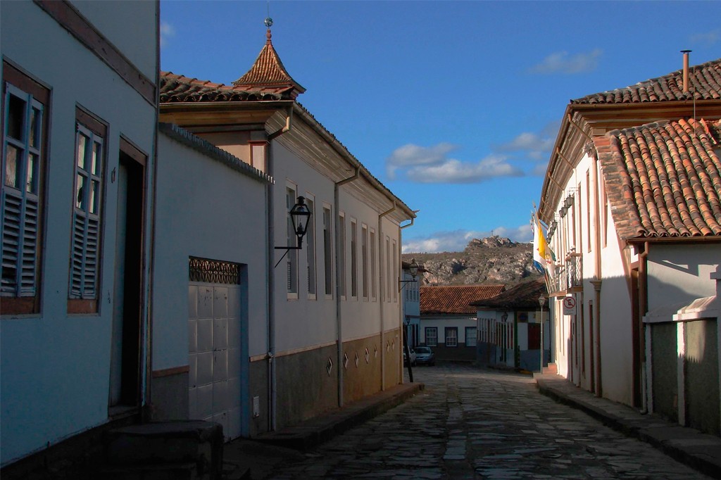 Rua de paralelep&iacute;pedos em cidade hist&oacute;rica, ladeada por casar&otilde;es coloniais de telhados de barro e paredes claras. &Agrave; esquerda, uma torre pontiaguda no telhado. Ao fundo, montanhas sob c&eacute;u azul com nuvens esparsas