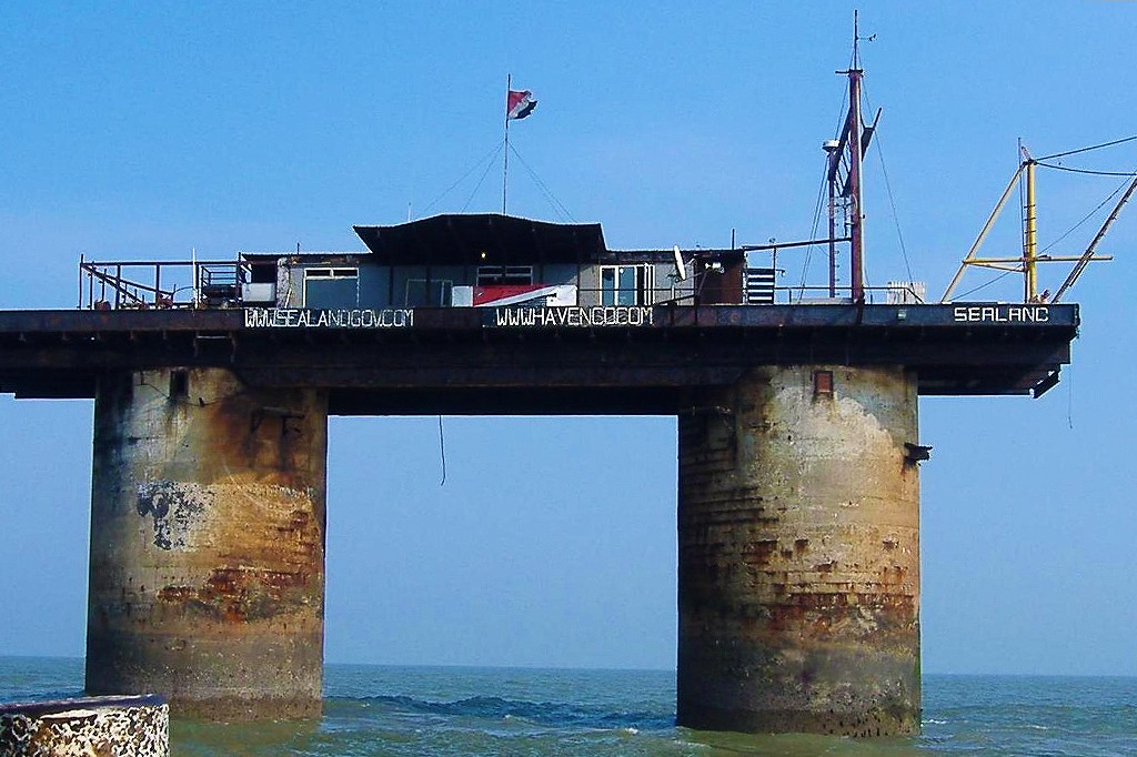 Plataforma marítima Sealand alguns meses após o incêndio, com estrutura de concreto e metal enferrujada, sustentando um edifício cinza com janelas e uma bandeira vermelha e preta. O mar azul-esverdeado rodeia a base, e o céu está claro. As palavras WWW.SEALANDGOV.COM, WWW.HAVENGB.COM e SEALAND estão visíveis na estrutura