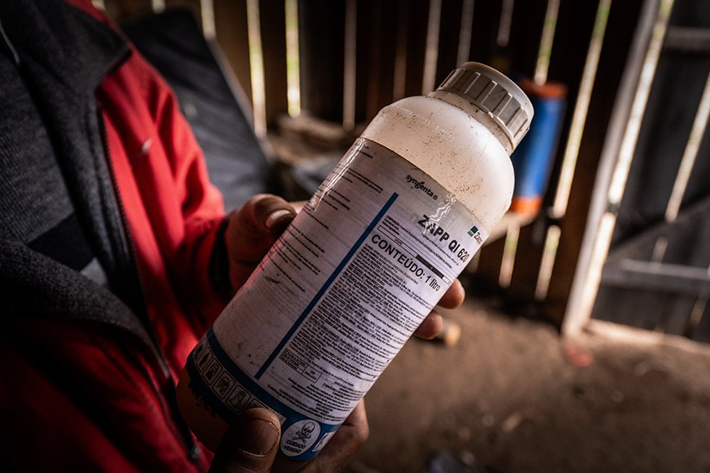M&atilde;o de uma pessoa com jaqueta vermelha e cinza segurando um frasco branco de agrot&oacute;xico Syngenta Zapp QI 620, com r&oacute;tulo azul e branco, que indica Conte&uacute;do: 1 litro e um s&iacute;mbolo de caveira com a frase Cuidado Veneno