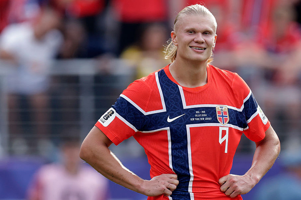 Jogador de futebol Erling Haaland sorrindo, com cabelo loiro preso, vestindo camisa vermelha da Noruega com cruz azul e branca, m&atilde;os na cintura, em campo de futebol.