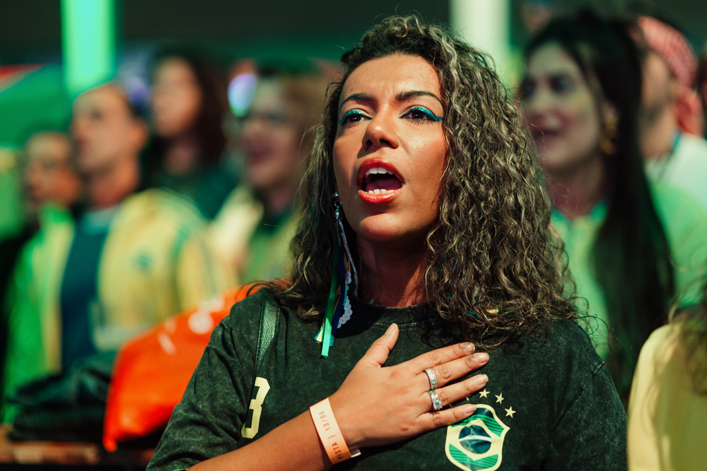 Mulher de cabelos cacheados, maquiagem azul e batom vermelho, canta com a mão no peito, vestindo uma camiseta verde escura com o escudo da seleção brasileira. Outras pessoas desfocadas aparecem ao fundo.