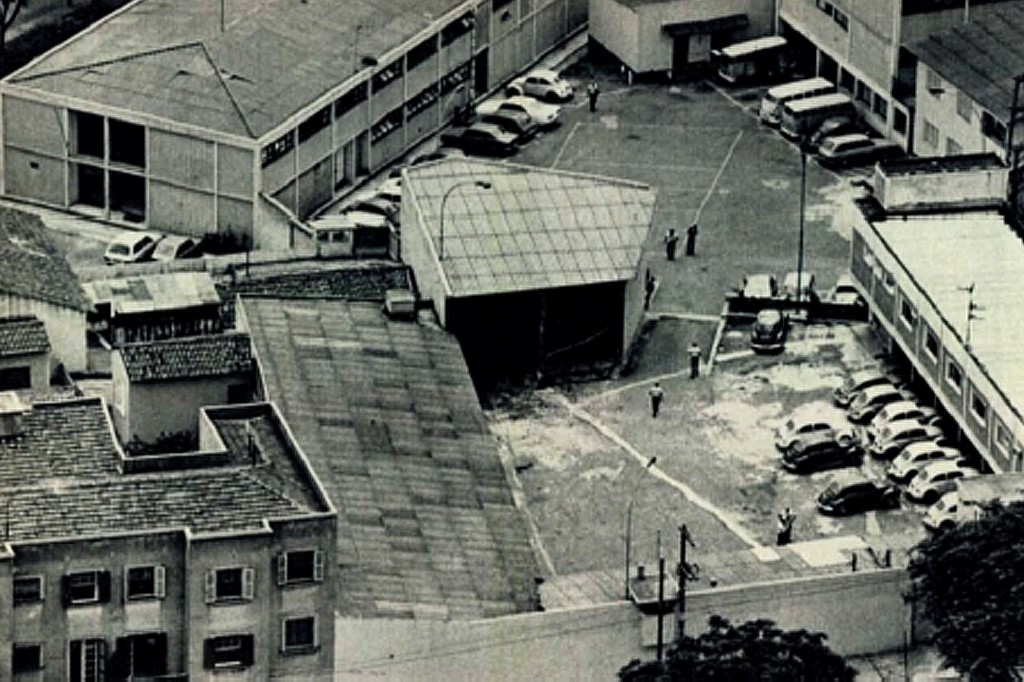 Vista aérea em preto e branco de um pátio com carros estacionados e pessoas caminhando, cercado por edifícios de diferentes tamanhos e telhados variados, com algumas árvores no canto inferior direito
