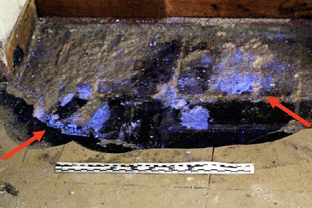 Manchas azul-violeta fluorescentes, indicando vest&iacute;gios de sangue, em um piso de concreto danificado, com duas setas vermelhas apontando para as &aacute;reas de maior concentra&ccedil;&atilde;o. Uma r&eacute;gua de escala branca e preta est&aacute; no ch&atilde;o, abaixo das manchas