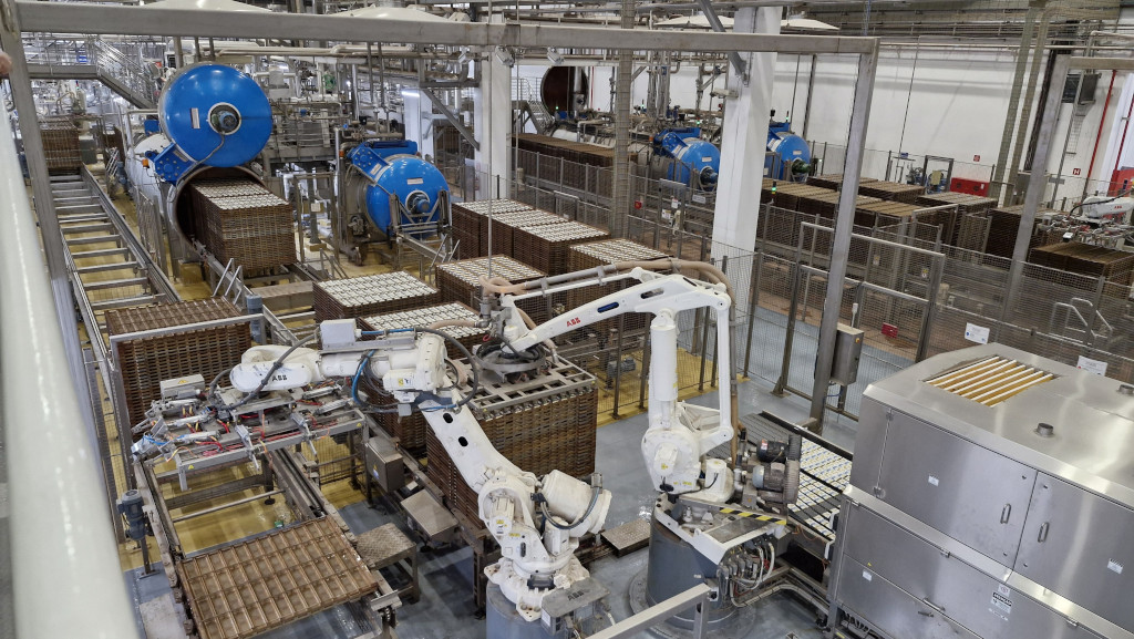 Visão aérea de uma fábrica automatizada com esteiras transportadoras, robôs brancos da marca ABB e grandes máquinas azuis cilíndricas, processando caixas e bandejas de produtos em um ambiente industrial limpo