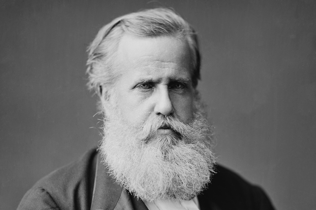 Retrato em preto e branco de um homem de meia-idade com barba e cabelo grisalhos, olhar sério e direto para frente, vestindo um terno escuro e camisa clara.