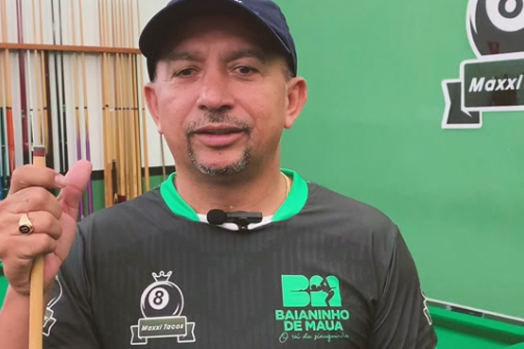 Homem de boné azul e camisa preta com detalhes verdes, segurando um taco de sinuca, com um microfone preso à gola. Ele tem barba rala e um anel dourado no dedo anelar. Ao fundo, um suporte com vários tacos e uma parede verde com um logo de sinuca.