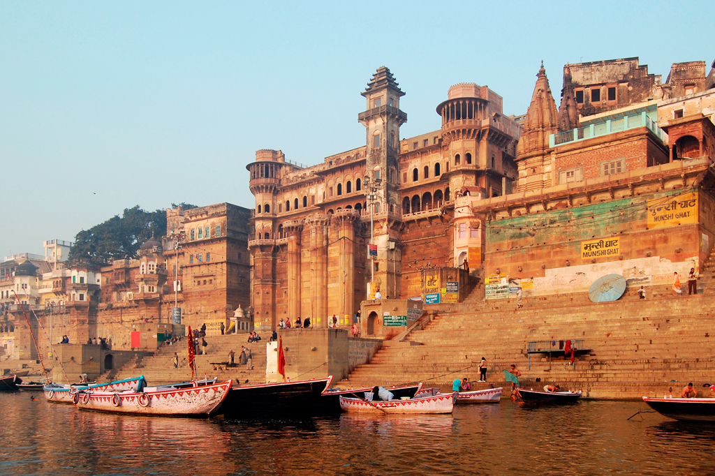 Barcos coloridos flutuam no rio Ganges, com edifícios históricos de pedra e escadarias em ghats de Varanasi, Índia. Pessoas caminham pelas escadas e interagem com a água sob um céu claro