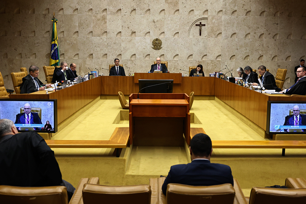 Sessão plenária do Supremo Tribunal Federal com ministros sentados em uma mesa semicircular de madeira, usando togas pretas, e dois homens assistindo de costas em primeiro plano. Ao fundo, uma parede de mármore com o brasão da República e uma cruz, e a bandeira do Brasil. Dois monitores exibem um ministro em videoconferência