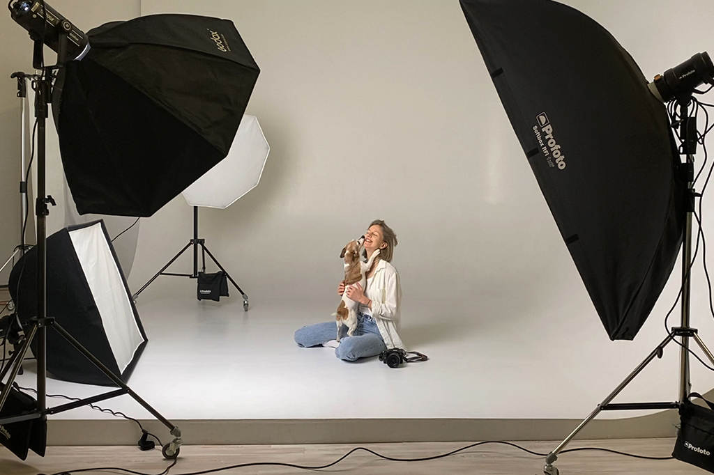 Mulher loira sorridente, de camisa branca e jeans, sentada no chão de um estúdio fotográfico branco, abraçando um cachorro marrom e branco que lambe seu rosto. Equipamentos de iluminação com softboxes pretos e brancos estão ao redor