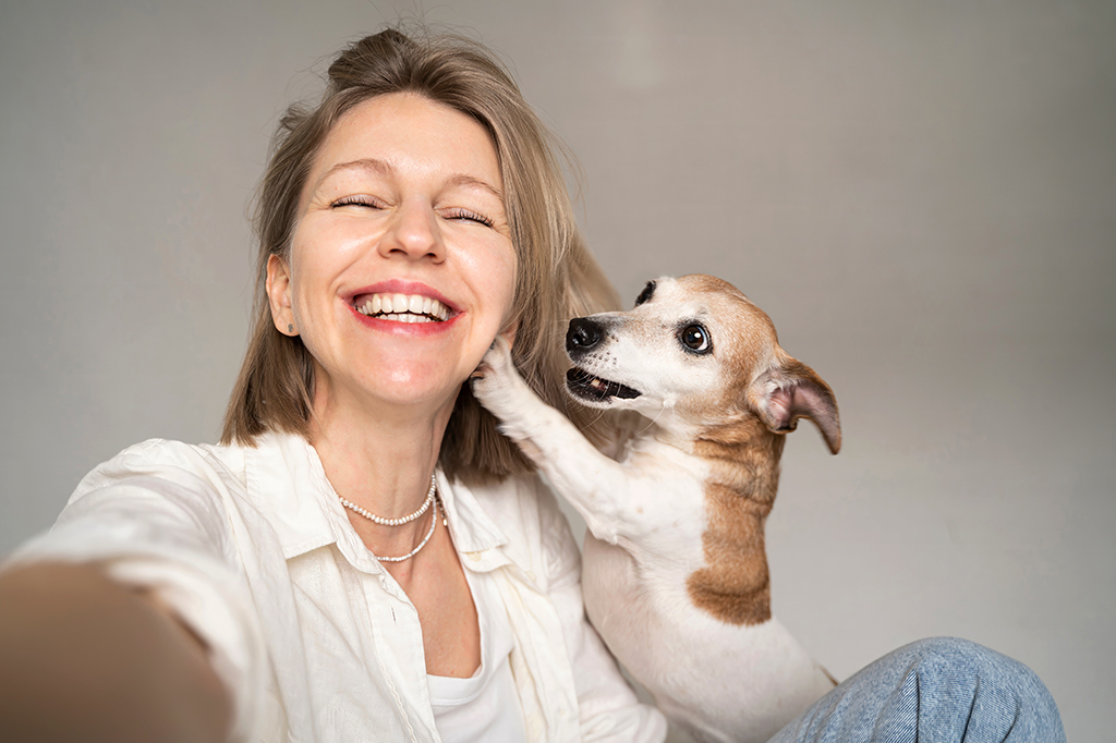 Mulher loira sorrindo com os olhos fechados, usando camisa branca e colar de pérolas, enquanto um cachorro marrom e branco com a pata na orelha dela a olha