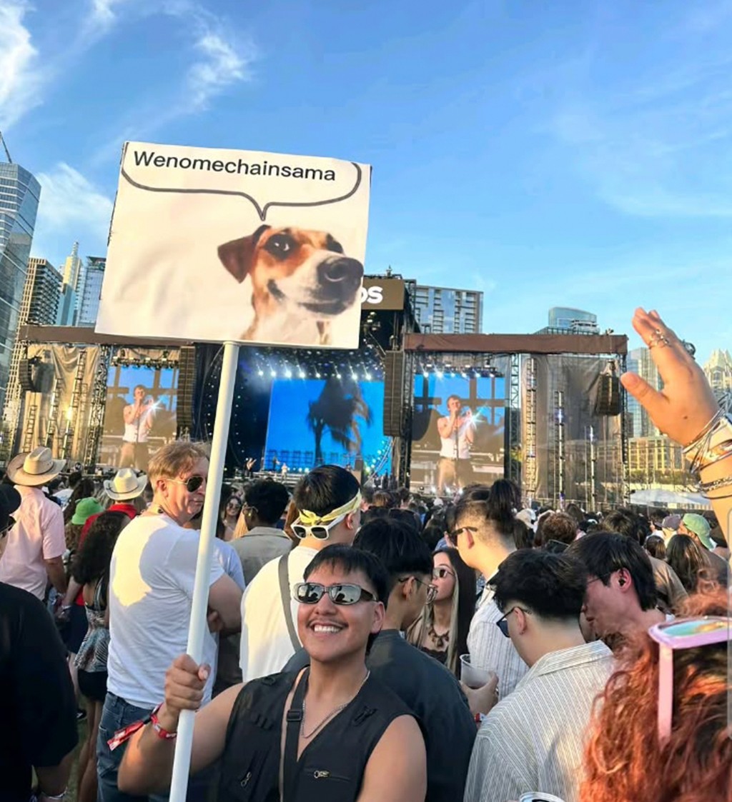 Homem sorridente de óculos escuros segura cartaz com foto de cachorro e balão de fala Wenomechainsama em show lotado, com palco e telões ao fundo exibindo um cantor