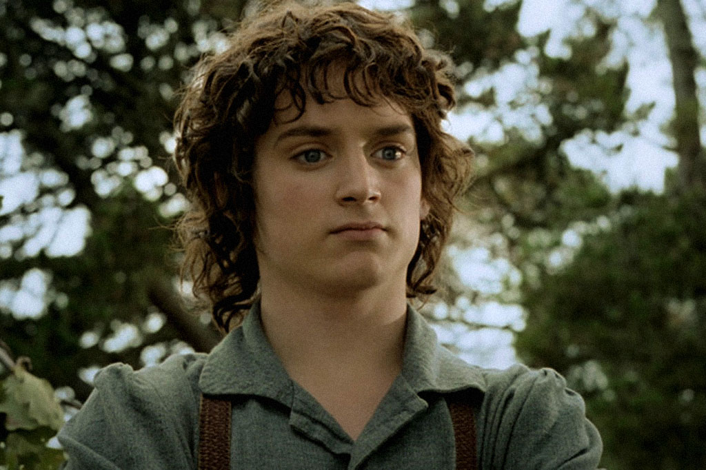 Close-up de Frodo Bolseiro, um jovem com cabelos castanhos encaracolados e olhos azuis, vestindo uma camisa verde-acinzentada e suspensórios marrons, com uma expressão séria. O fundo desfocado mostra folhagem verde e pontos de luz