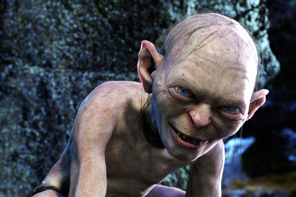 Gollum, criatura de pele pálida e enrugada, olhos azuis arregalados e dentes pontiagudos, sorri maliciosamente. Ele está curvado, com o corpo magro e ombros à mostra, em um ambiente escuro e rochoso