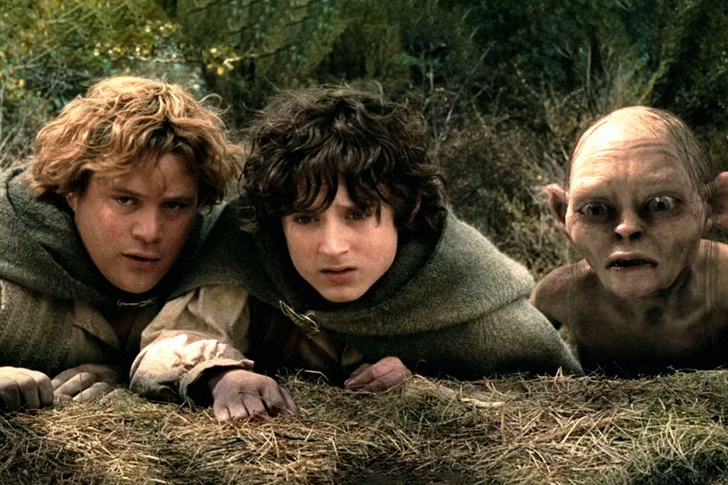 Samwise, Frodo e Gollum, personagens de O Senhor dos Anéis, deitados na grama seca, com expressões de preocupação e medo. Samwise e Frodo usam capas verdes, enquanto Gollum, à direita, tem pele pálida e olhos arregalados