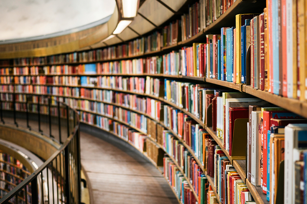 Prateleiras de madeira curvadas repletas de livros coloridos em uma biblioteca, com um corredor estreito e um corrimão preto à esquerda