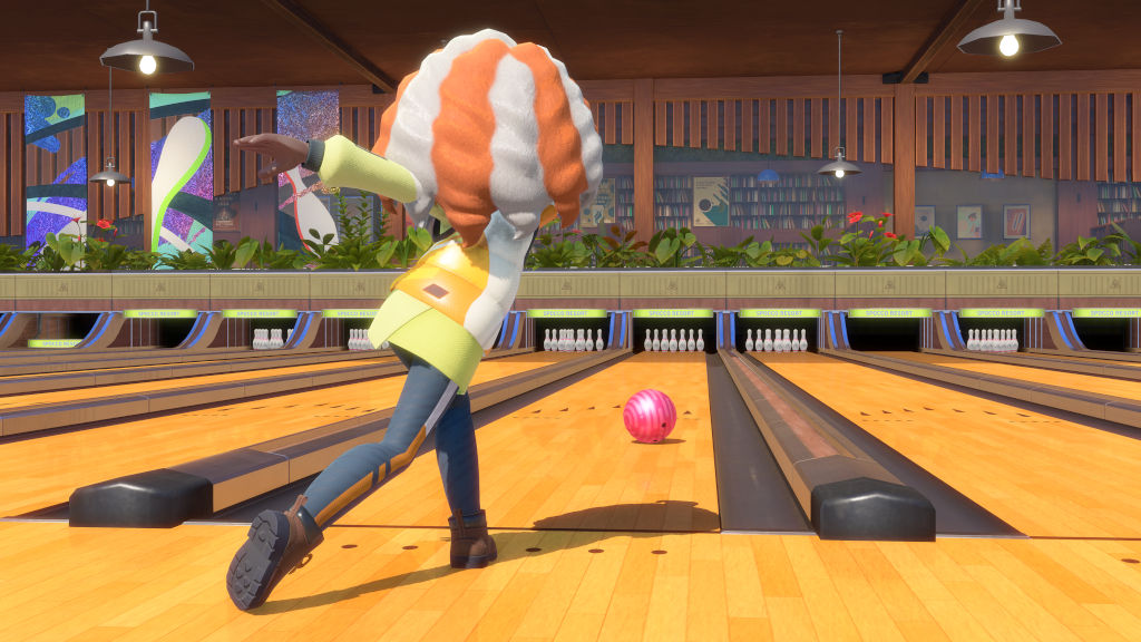 Personagem de jogo eletrônico com cabelo laranja e branco, camisa amarela e calça azul, arremessando uma bola de boliche rosa em uma pista de madeira clara. Ao fundo, outras pistas com pinos, plantas e estantes de livros