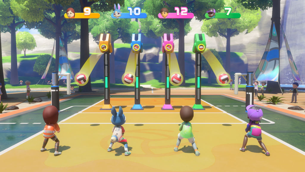 Quatro personagens de videogame, incluindo um coelho azul, jogam vôlei em uma quadra amarela e azul. Bolas de vôlei vermelhas e brancas caem de tubos coloridos, enquanto placares flutuam acima com números 9, 10, 12 e 7. O cenário tem árvores e uma tenda colorida