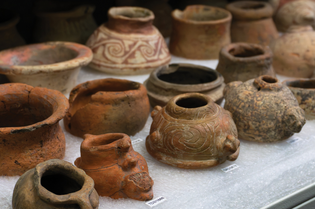 Vários vasos e potes de cerâmica antigos, em tons terrosos, alguns com padrões espirais e geométricos, exibidos sobre uma superfície branca com bolhas de ar para proteção, com etiquetas de identificação visíveis