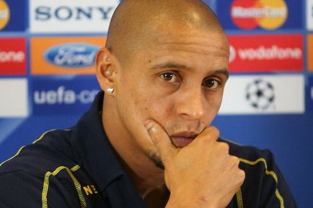 O jogador de futebol Roberto Carlos, careca, com brinco de coração na orelha esquerda, veste camisa azul-marinho com detalhes amarelos, pensativo, com a mão direita no queixo, em frente a um painel com logotipos de patrocinadores como Sony, Ford e UEFA Champions League