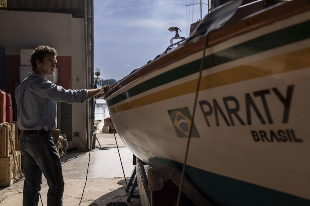 Homem de camisa azul e calça jeans toca um barco branco com faixas verde e amarela, onde se lê PARATY BRASIL e a bandeira brasileira, em uma doca ensolarada