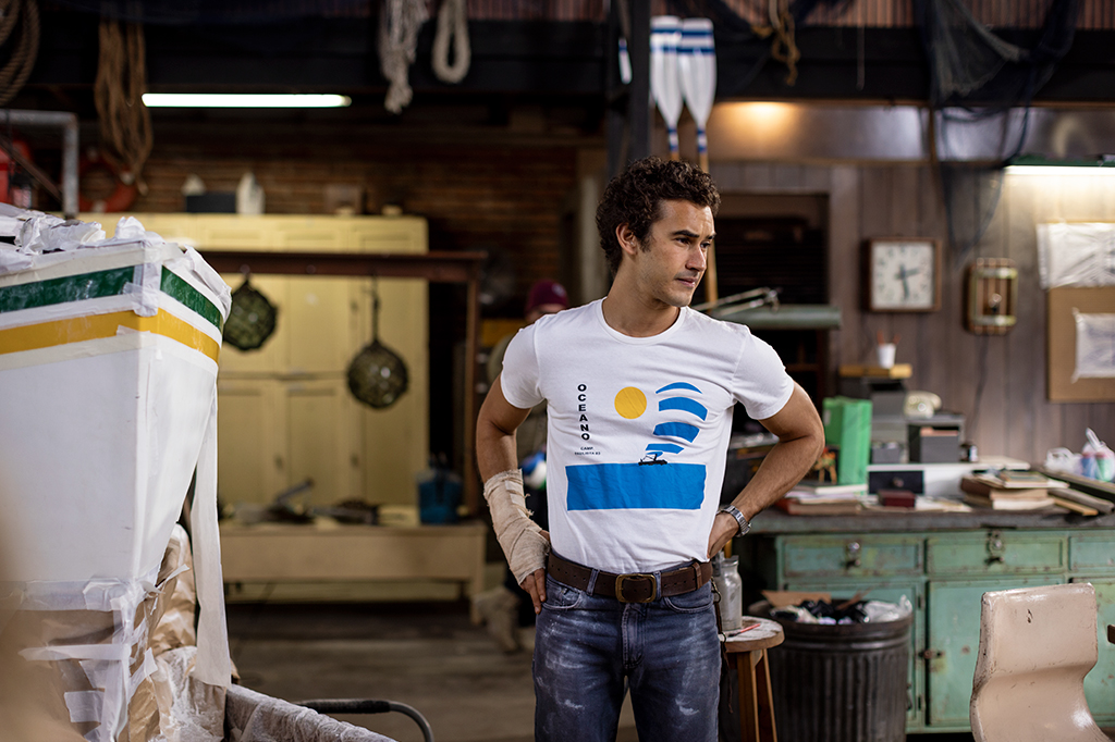 Homem jovem de cabelo encaracolado, camiseta branca com estampa de veleiro e sol, jeans e cinto marrom, com atadura no antebraço esquerdo, em uma oficina de barcos com ferramentas e redes ao fundo