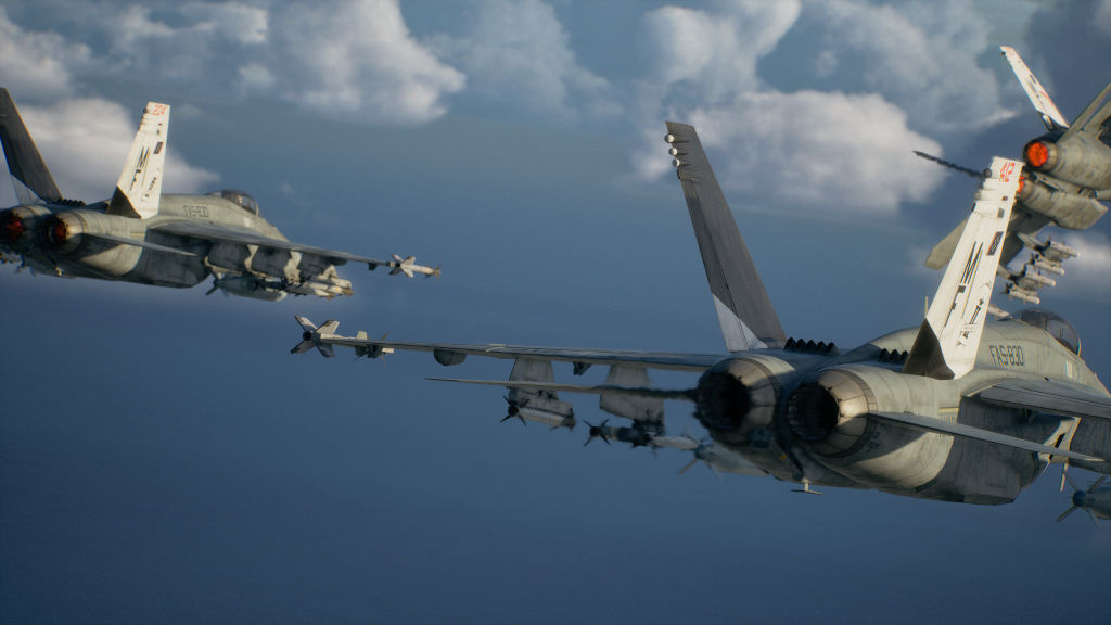 Três caças militares cinzas voam em formação contra um céu azul com nuvens brancas. Dois aviões estão em primeiro plano, um à esquerda e outro à direita, com seus motores e asas visíveis. O terceiro avião está mais distante, à direita, com a cauda e parte da asa visíveis. Todos os aviões estão equipados com mísseis sob as asas