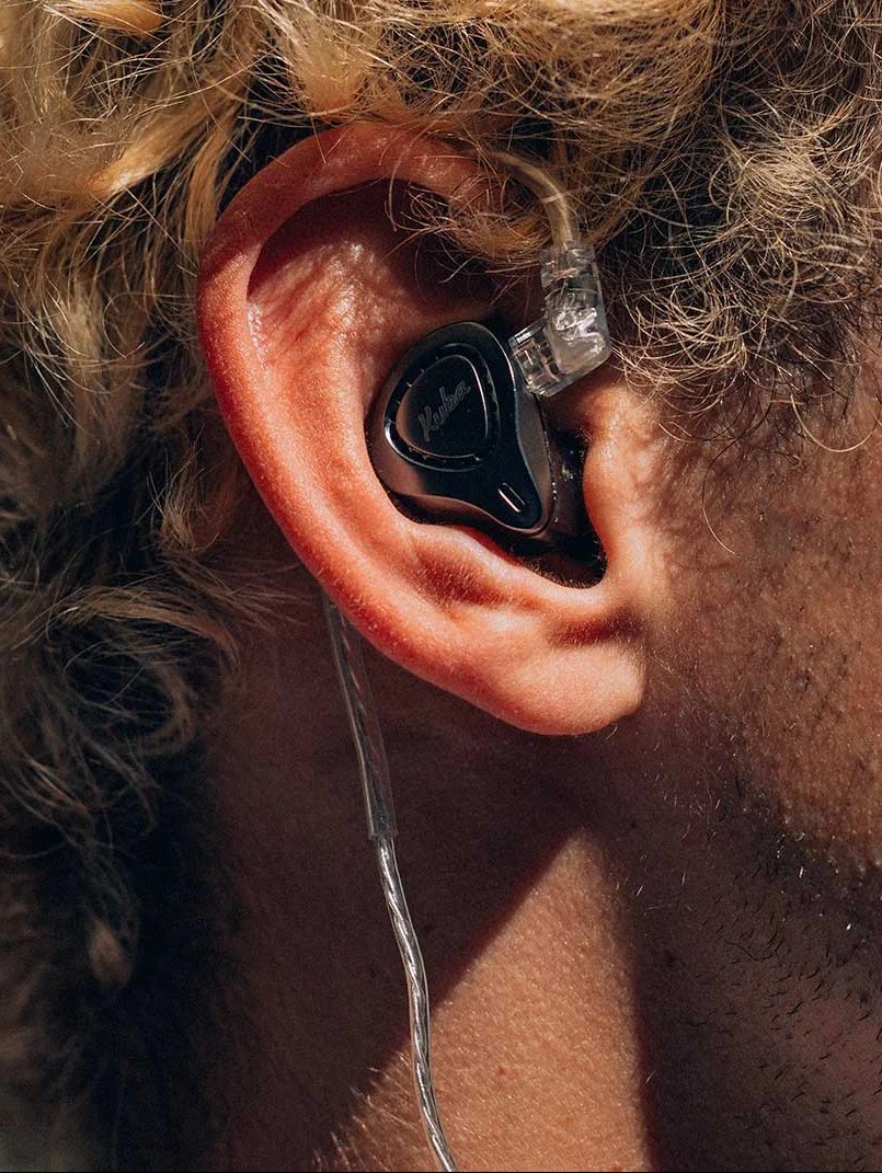 Close-up do lado direito do rosto de uma pessoa com cabelo loiro encaracolado, usando um fone de ouvido intra-auricular preto com o nome Kuba em branco, conectado por um cabo transparente que passa por trás da orelha